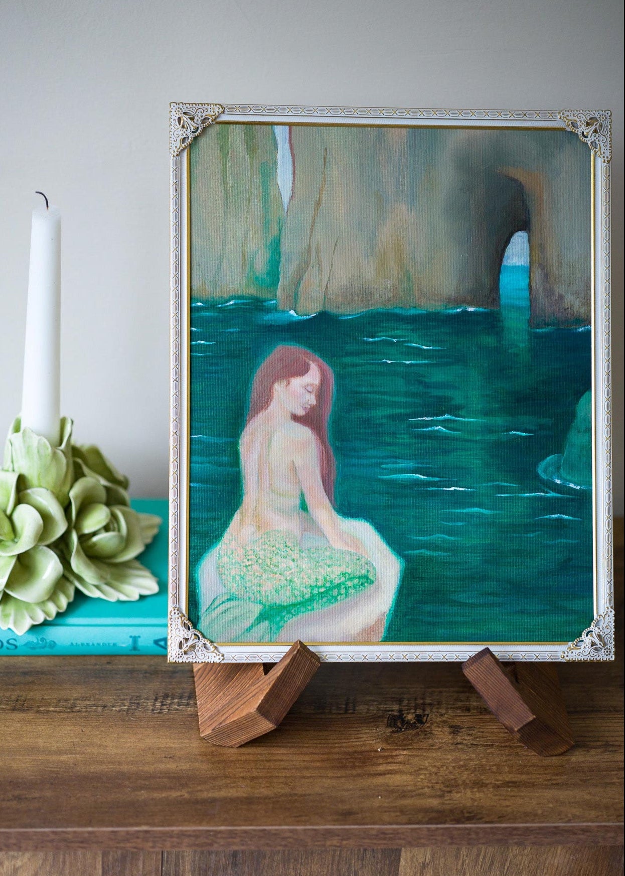 A framed painting of a mermaid in a sea cave, displayed on a wooden stand on a table next to a teal book, a white candle, and a green flower arrangement.